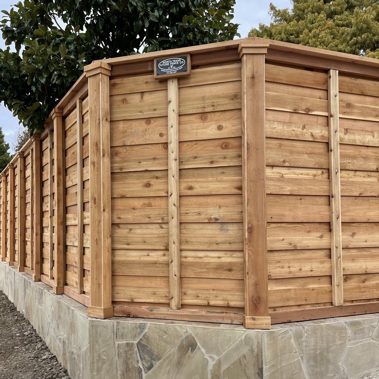 A finished cedar fence corner with cap, trim and boxed posts on a flagstone base wall, carrying the North Texas Future Fence cast oval plaque