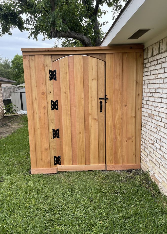 An arched-top cedar gate with black strap hinges against a white brick wall