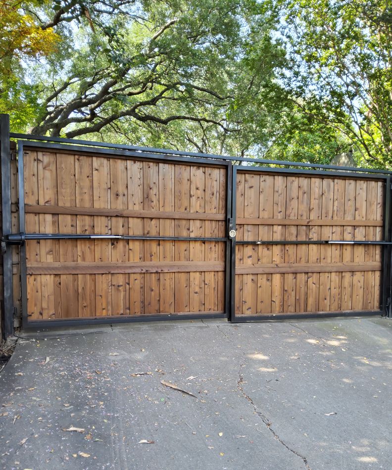 A wide double cedar driveway gate on a black steel frame, closed across a residential drive