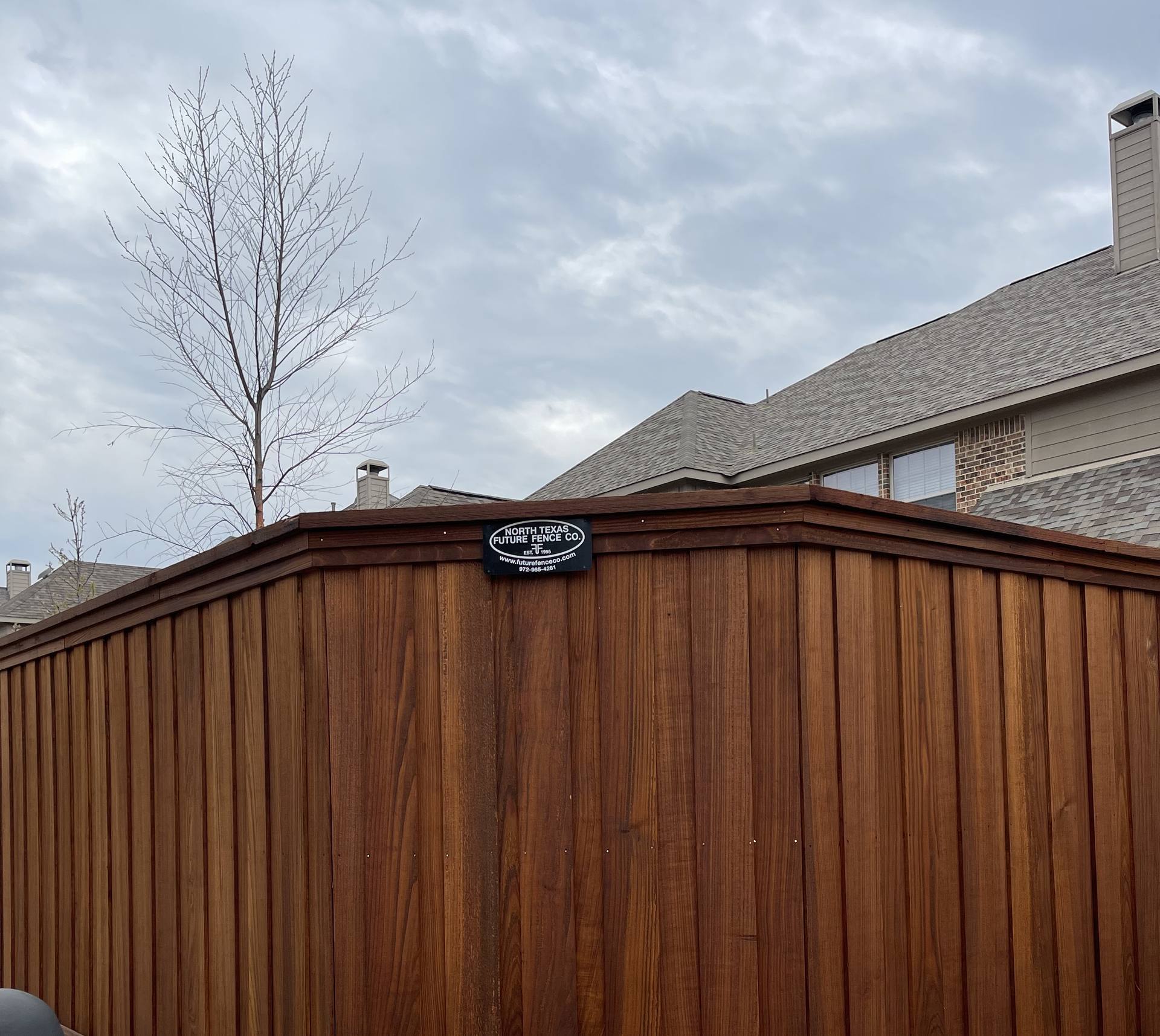An espresso-stained scallop-top fence on a white stone base with the Future Fence builder plaque