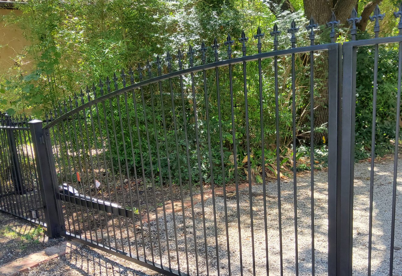 An arched black wrought-iron double gate with fleur-de-lis finials across a gravel drive