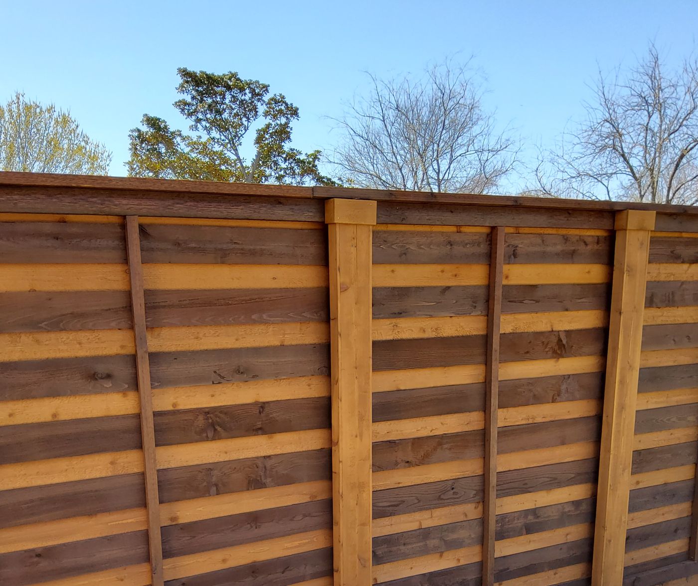 A two-tone horizontal board-on-board fence with fresh stain under a clear blue sky