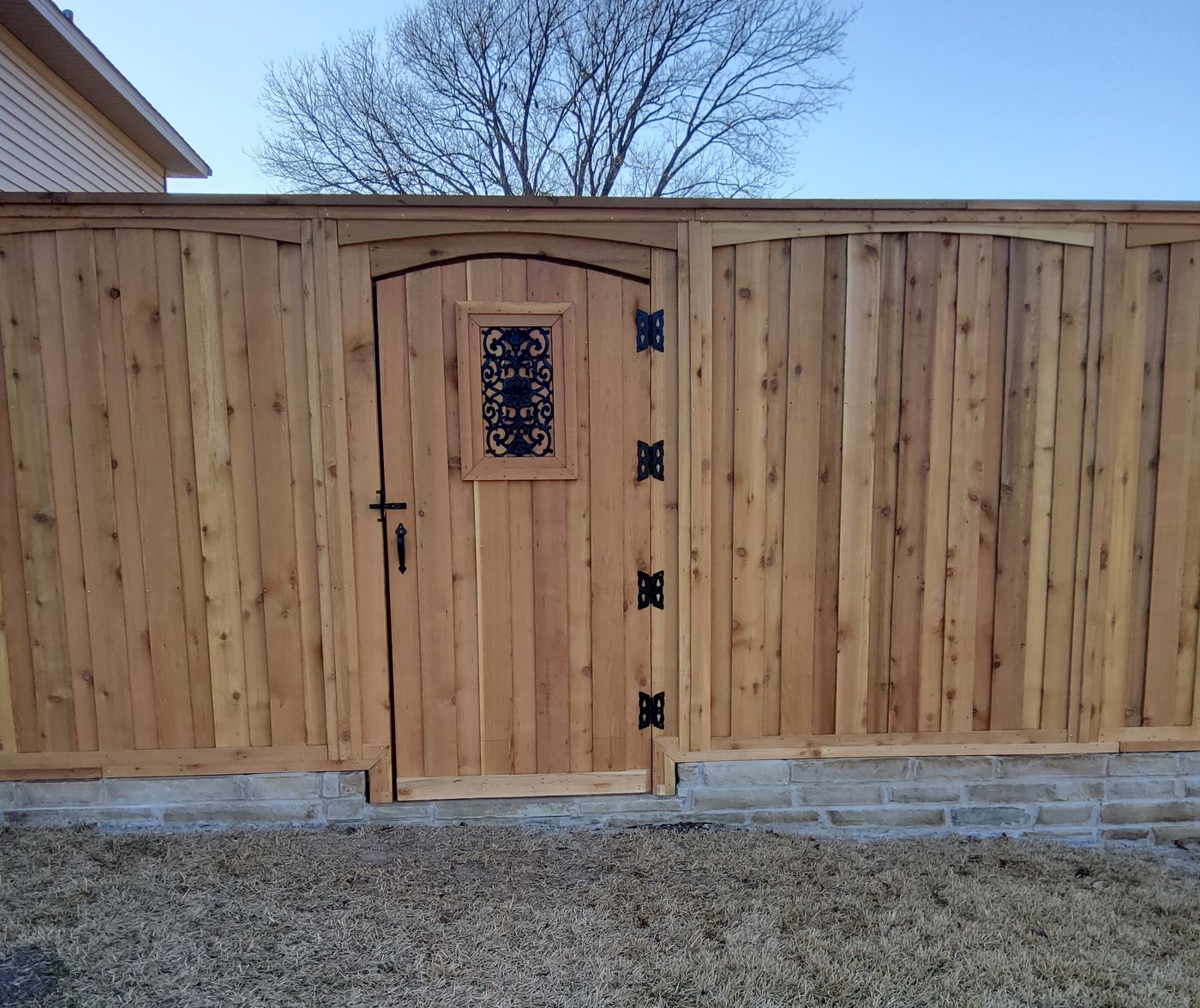 An arched cedar gate with a black iron scroll inset, flanked by scalloped board-on-board fence panels on a stone base