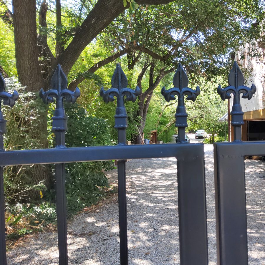 Black iron estate gates with fleur-de-lis finials across a gravel drive under a North Dallas oak canopy